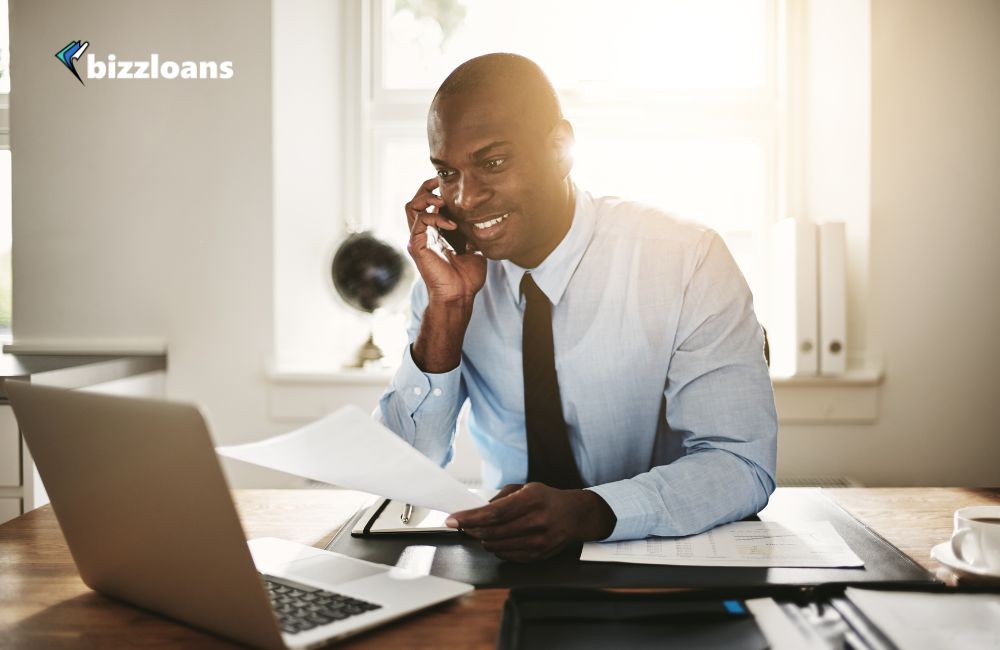 small-business-loan-brokers-do-you-need-one male broker talking to a client on the phone while holding a paper in front of laptop