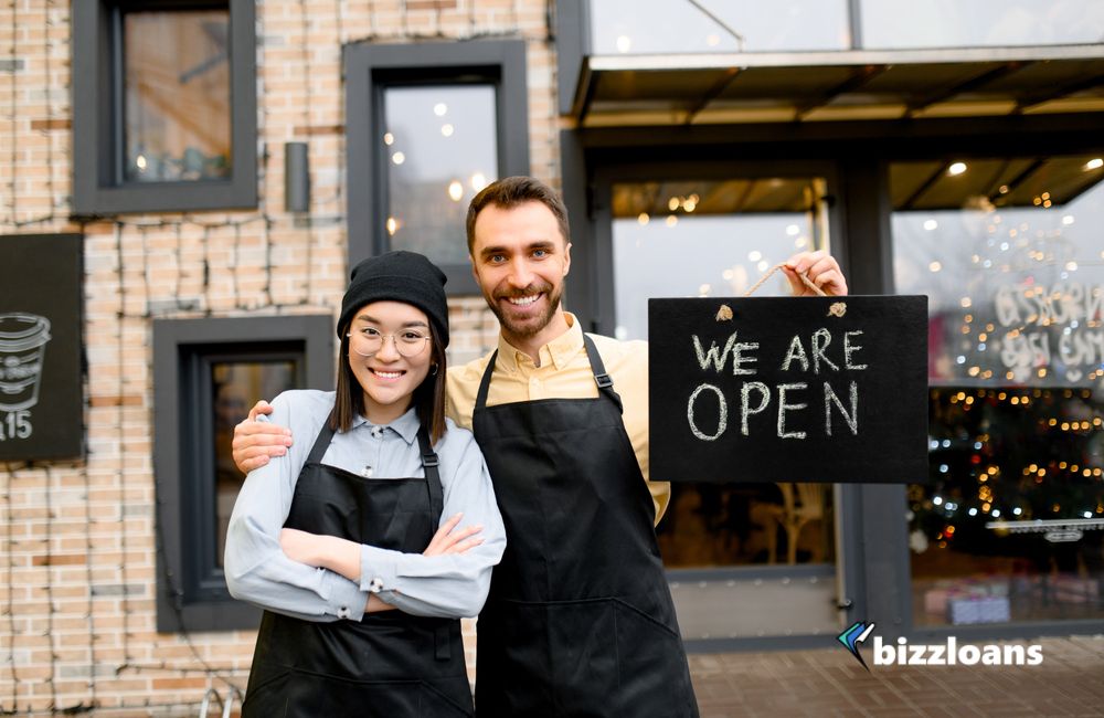 business-overdraft-and-its-benefits-for-your-business Two multiracial friendly waiters wearing uniform stand outside a restaurant, cafe or bar, showing signboard "We are OPEN", with pleasured smiling, welcome.